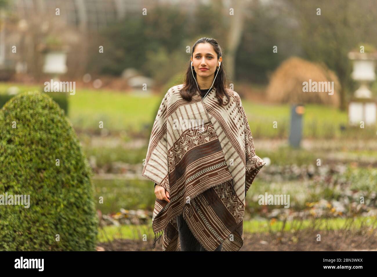 Femme indienne portant un poncho et écoutant des écouteurs debout dans un jardin topiaire ornemental. Banque D'Images