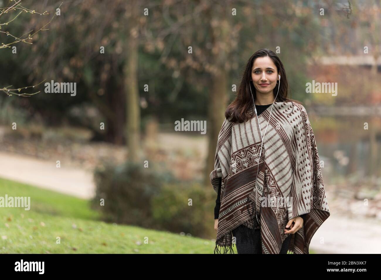 Jeune femme du Moyen-Orient portant un poncho à motifs et écoutant des écouteurs dans le parc. Banque D'Images