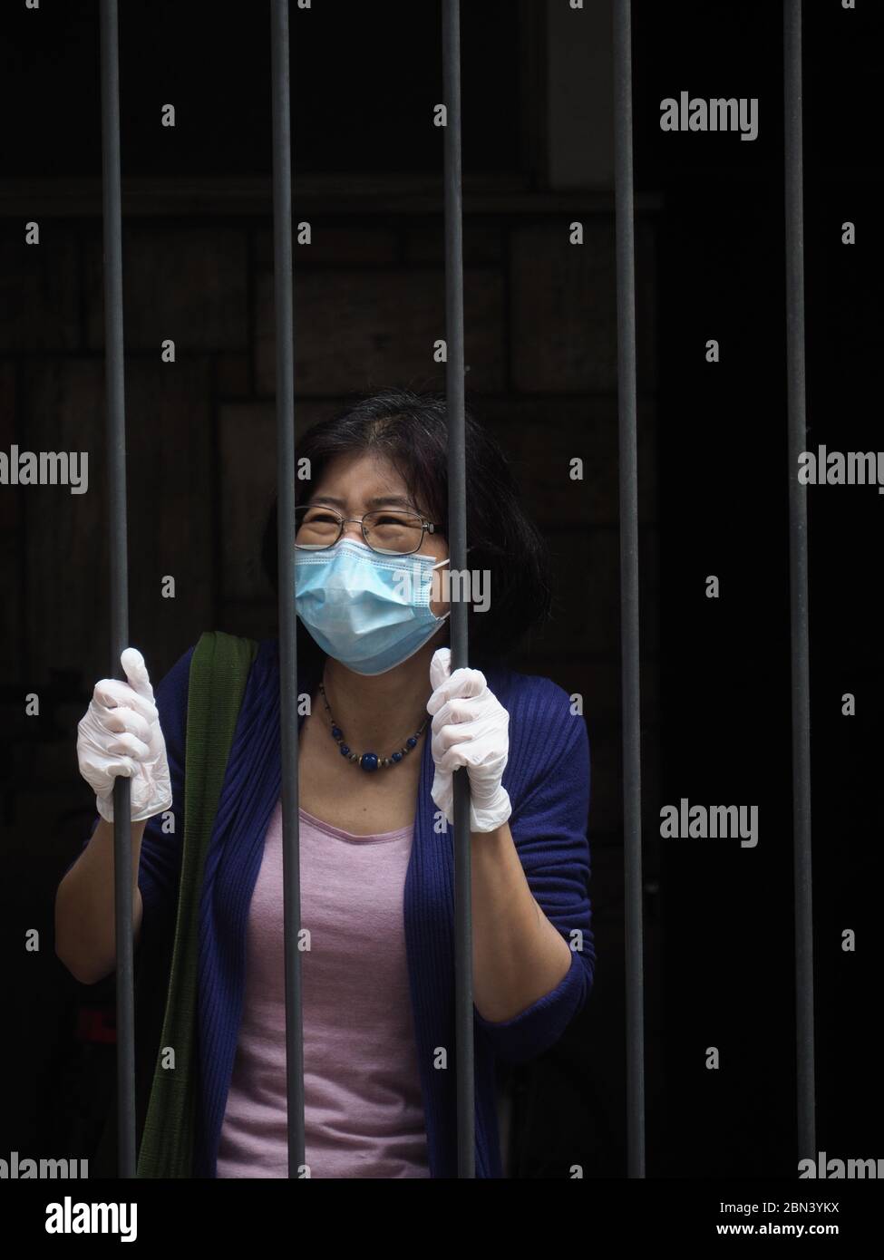 Femme asiatique avec masque et gants derrière une grille de barres - grille - pendant le confinement du coronavirus de confinement de la maladie COVID-19. Banque D'Images