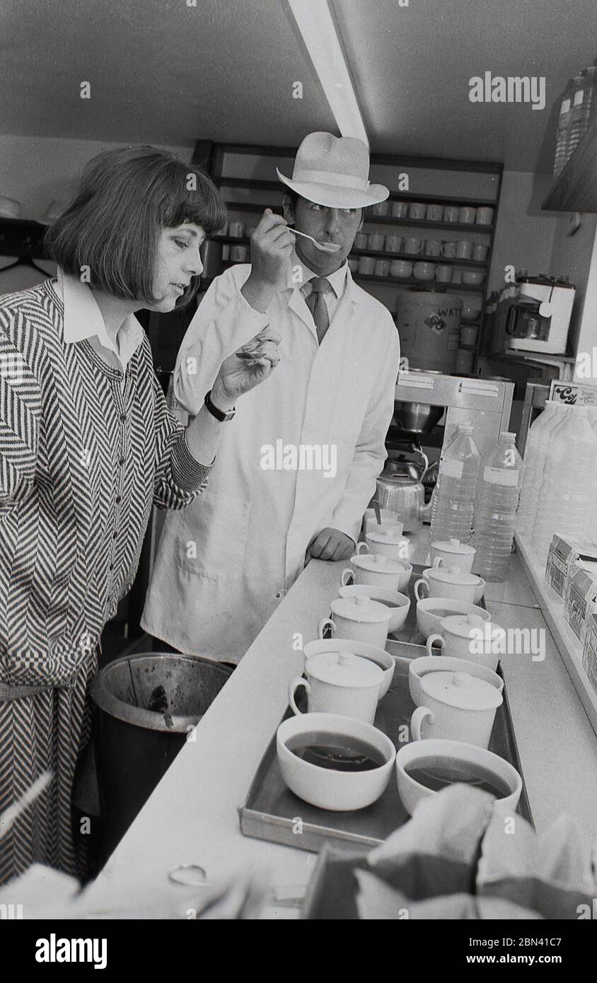 années 1980, dégustation de thé.. Un homme portant un manteau blanc et un chapeau, un dégustateur entraîné, debout à côté d'une femme, tous deux utilisant des cuillères pour goûter du thé, Harrogate, Angleterre, Royaume-Uni. Le thé, la plante Camelia Sinensis, a des saveurs et une apparence différentes de ses conditions de croissance et de ses procédés de fabrication. Banque D'Images