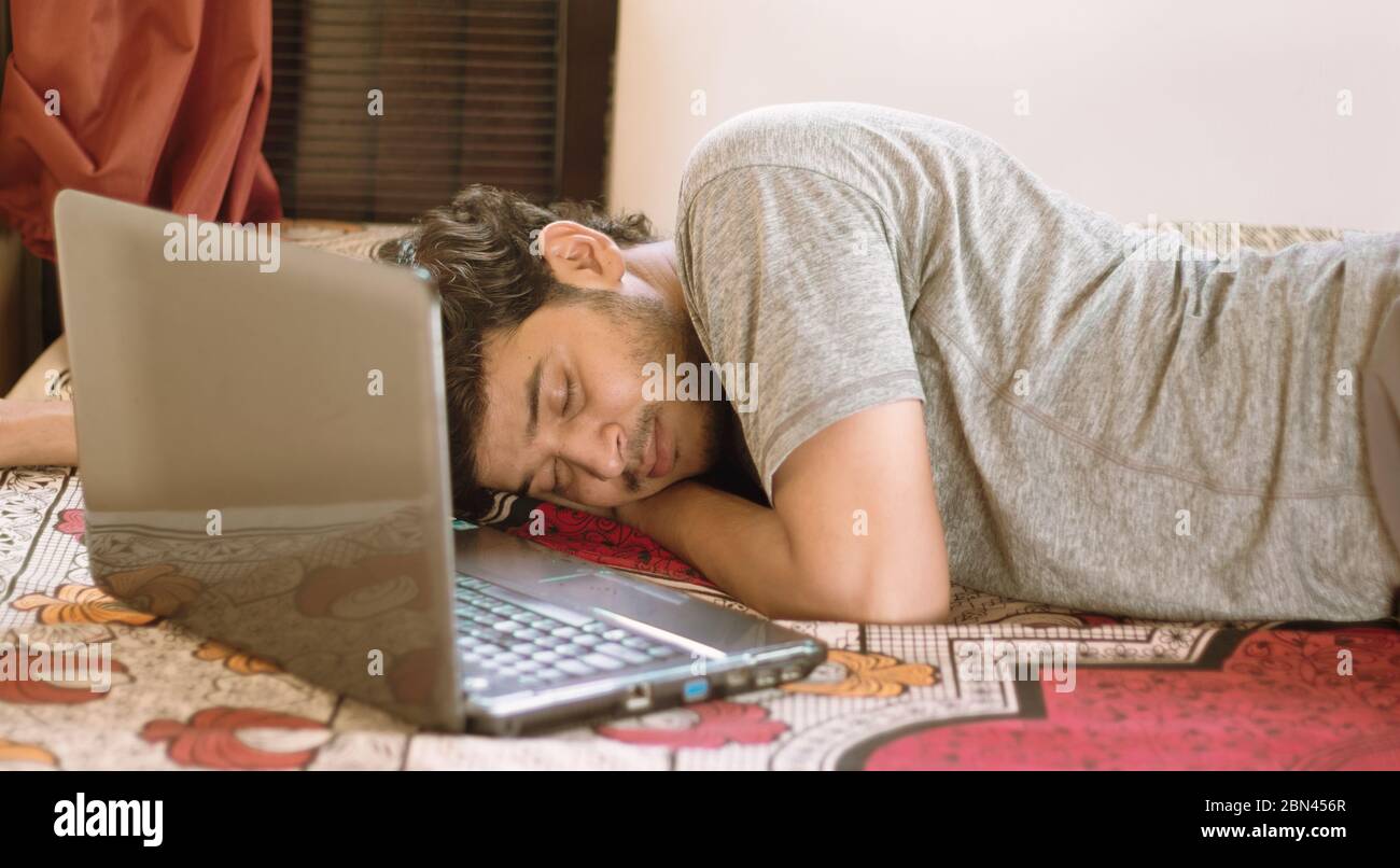 Jeune homme fatigué dormant au lit avec ordinateur portable - travail dur, paresse de travailler de la maison ou WFH pendant coronavirus ou covid-19 verrouiller le concept. Banque D'Images