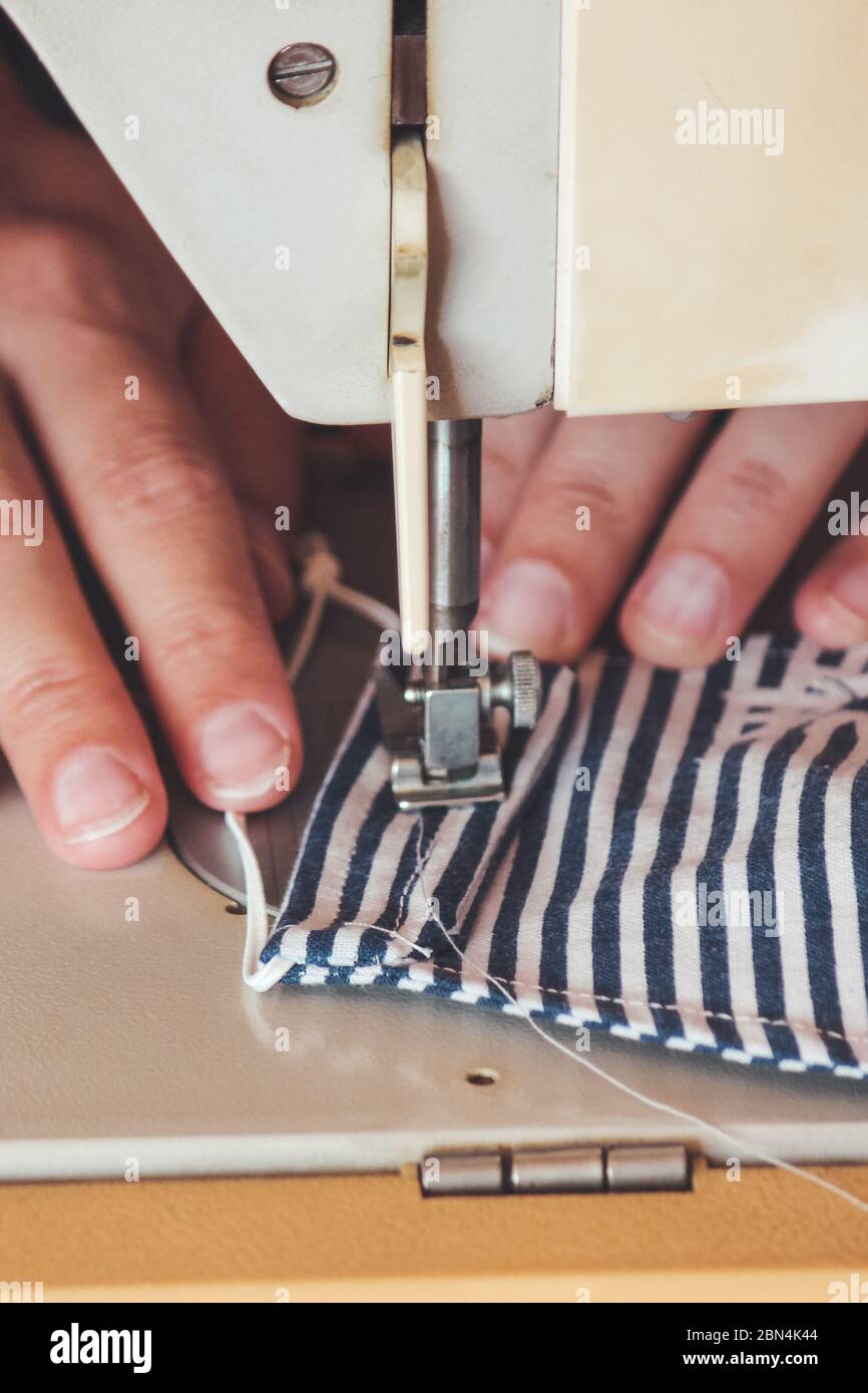 Les mains des femmes utilisant la machine à coudre pour coudre un masque médical coloré du visage pendant la pandémie de coronavirus. Masque de protection fait maison contre le virus. Masques à coudre, COVID-19. Mise au point sélective. Banque D'Images