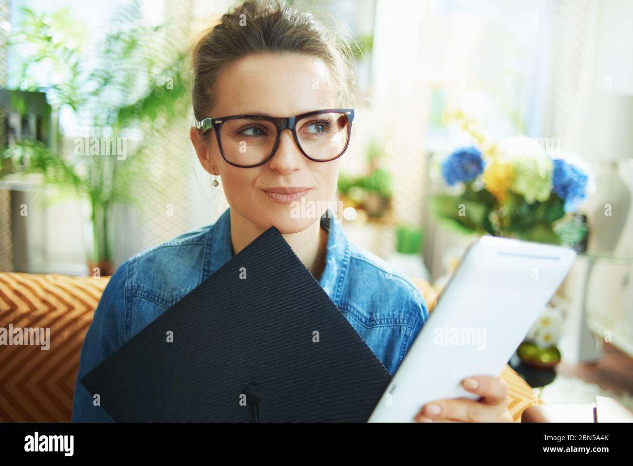 Jeune femme pensive en Jean avec capuchon de graduation et PC tablette à la maison moderne par temps ensoleillé. Banque D'Images