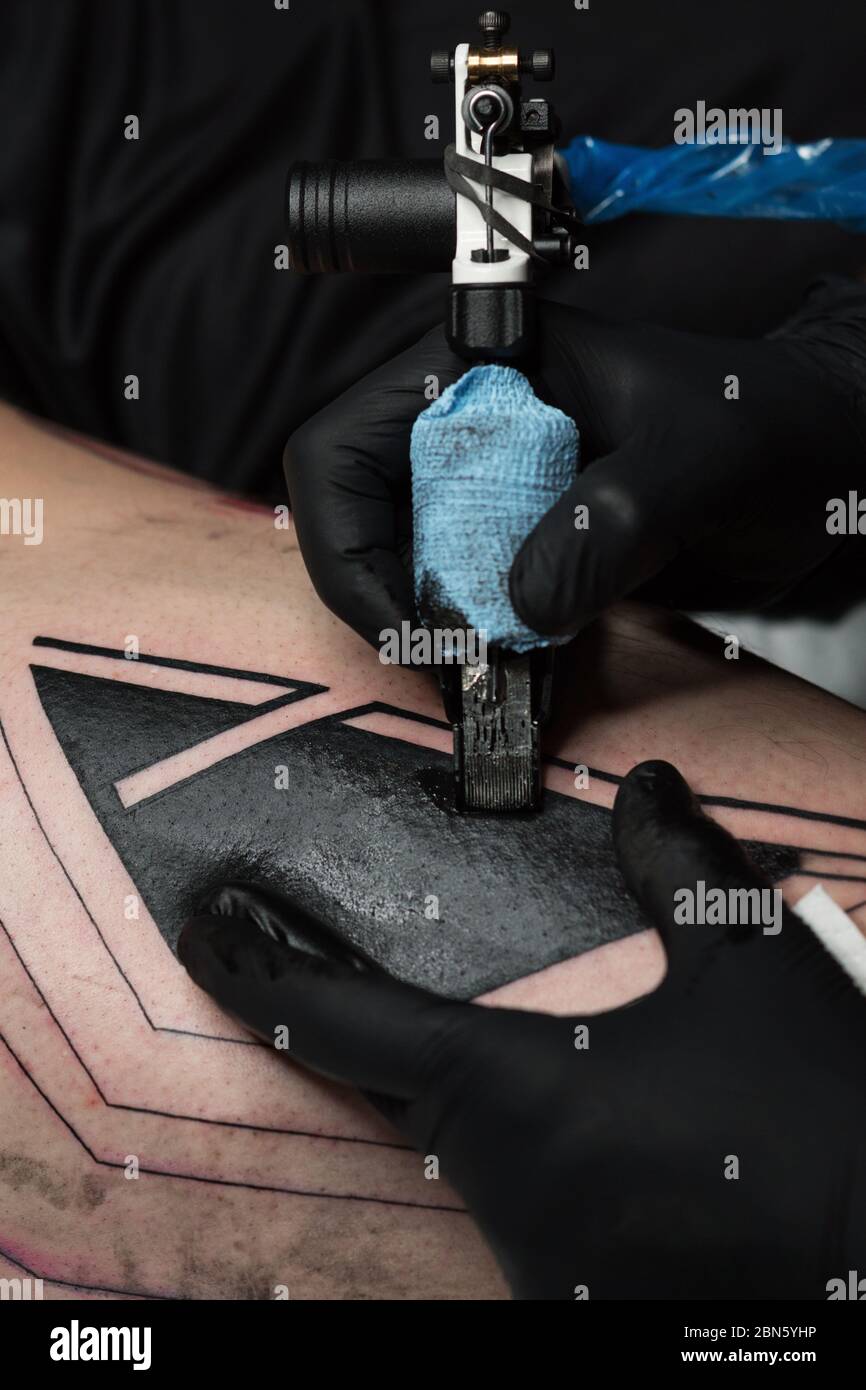 Un artiste professionnel de tatouage fait du tatouage sur la jambe de l'homme. Processus. Introduit l'encre noire dans la peau à l'aide de l'aiguille de la machine de tatouage. Gros plan sur la machine à tatouage. Tatouage. Gabarit de création d'image sur la jambe. Banque D'Images