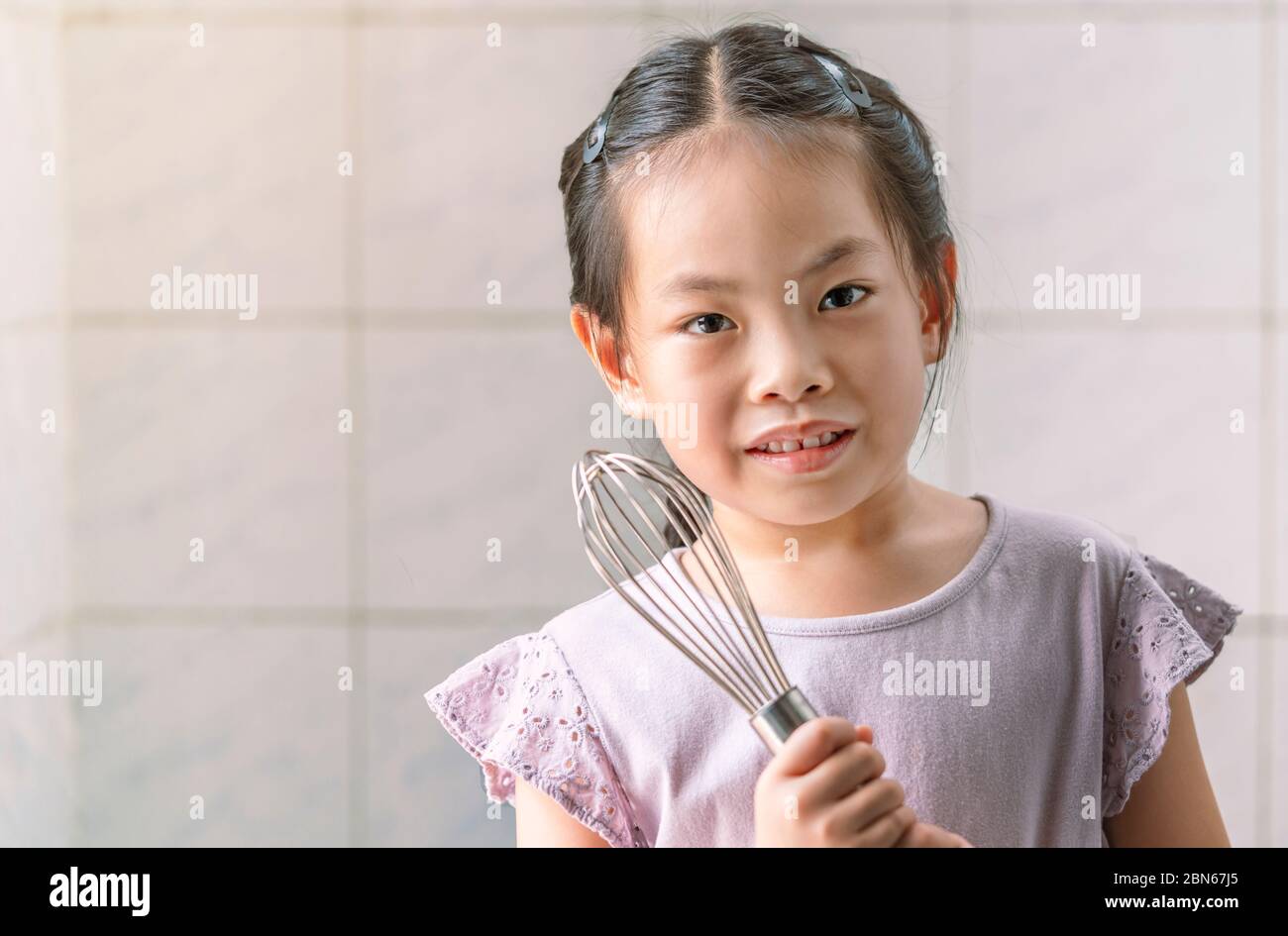 Portrait d'une fille asiatique tenant des ustensiles de cuisine ou un fouet à la main. Yeux regardant l'appareil photo. Enfant fille apprendre à cuisiner dans la cuisine. Cuisine de fond wa Banque D'Images