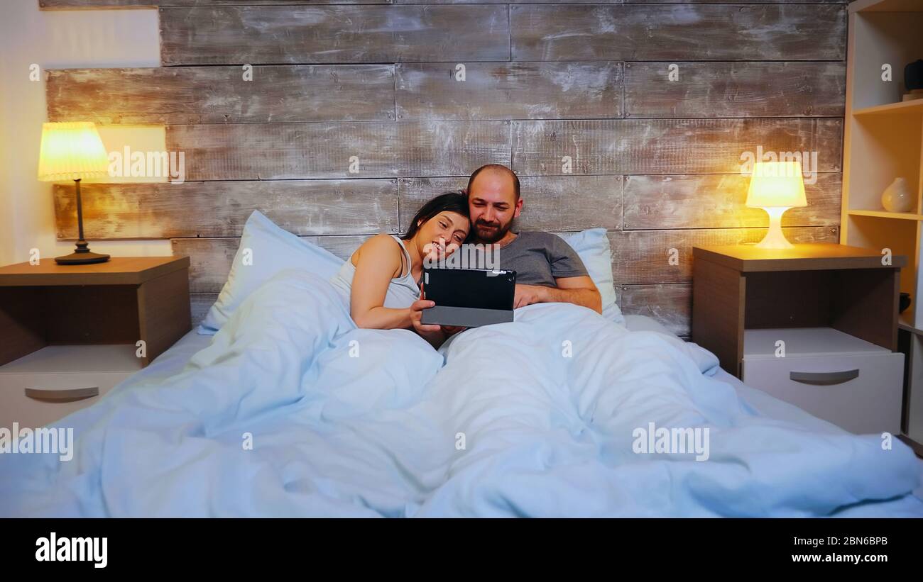 Zoom avant sur un jeune couple allongé dans le lit à l'aide d'une tablette avec ordinateur portant un pyjama. Banque D'Images