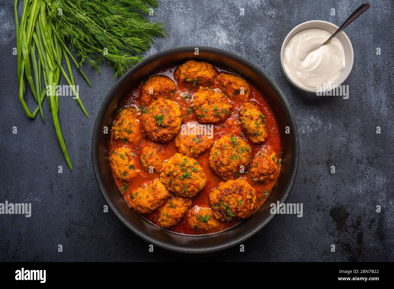 Boulettes de viande faites maison dans une sauce tomate dans une casserole sur fond sombre. À côté de la soucoupe avec de la crème aigre et des verts Banque D'Images