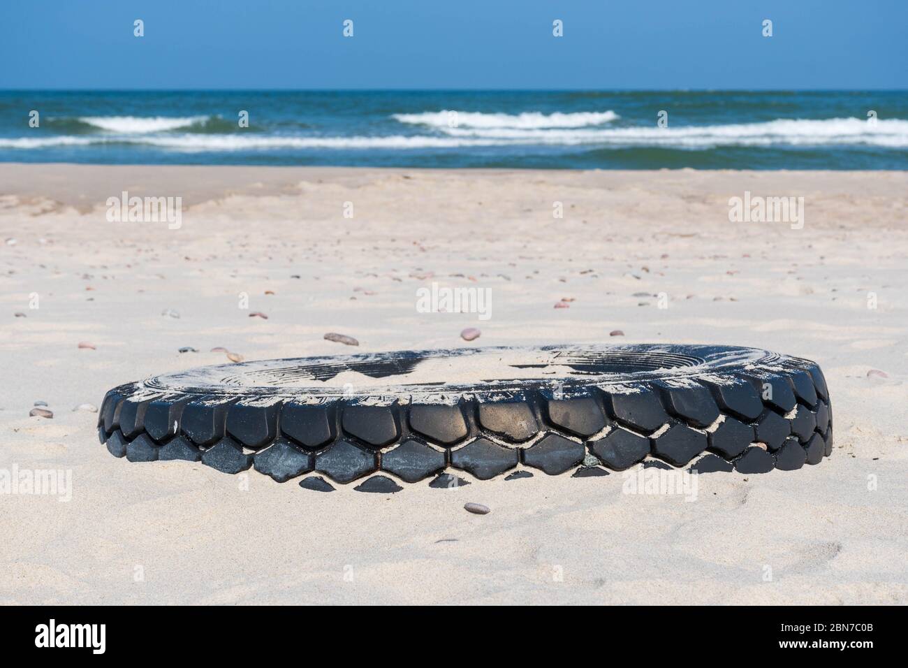Grand pneu en caoutchouc noir laissé sur une plage de sable avec mer bleue et vagues sur fond, environnement pollution concept Banque D'Images