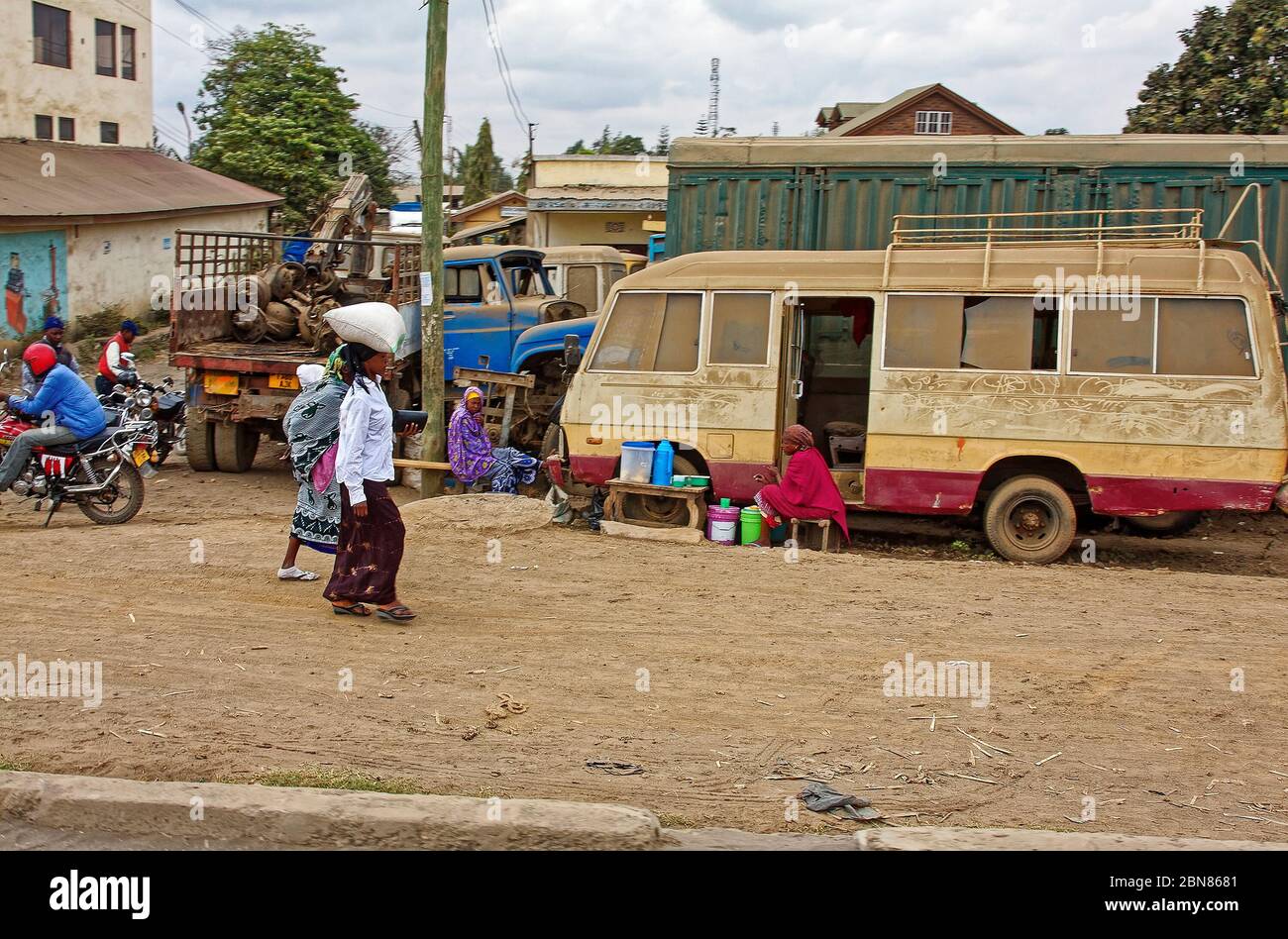 scène de rue, deux femmes marchant, sac équilibré sur la tête, femmes assises, petit vieux bus logements, hommes sur les motos, rue de terre, Afrique; Arusha; Tanzanie Banque D'Images