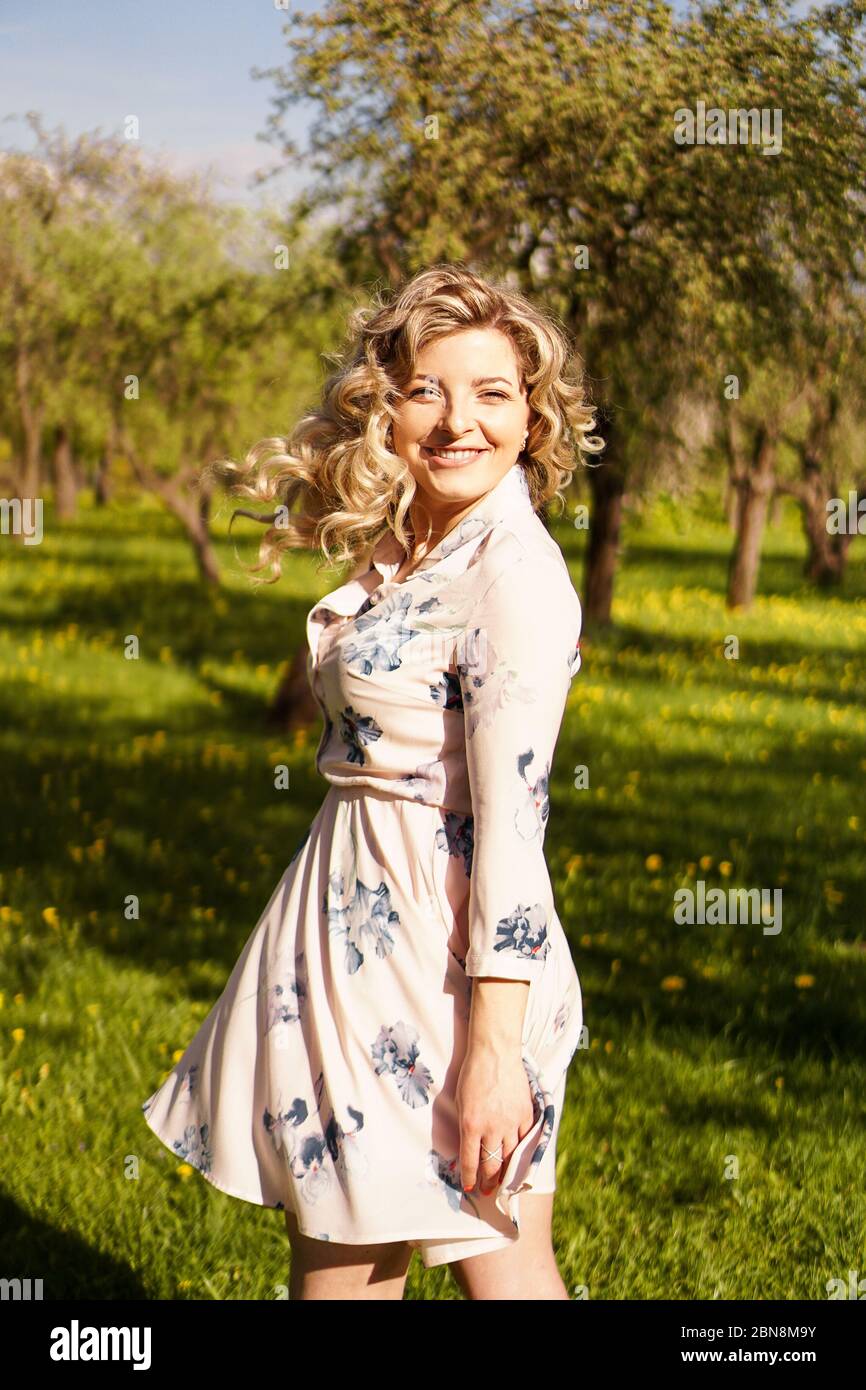 Bonne jeune femme aux cheveux blonds, portant une robe, posant à l'extérieur dans un jardin avec des cerisiers au soleil, souriant. Boucles qui flottent dans le vent Banque D'Images