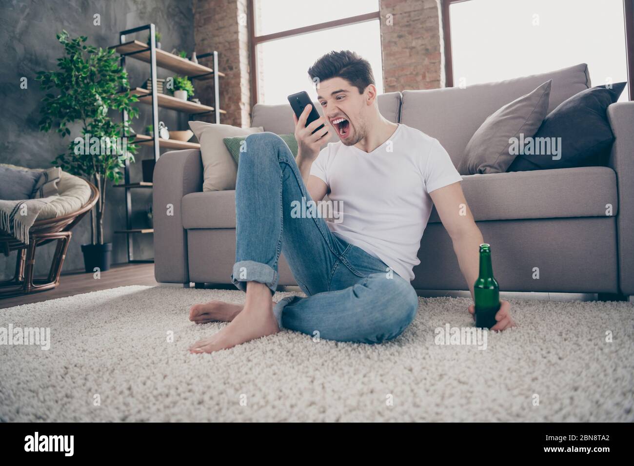 Portrait de son beau attirant fou fou fou frustré gars assis sur la moquette hurler sur le téléphone boire de la bière à l'intérieur moderne industriel loft brique Banque D'Images