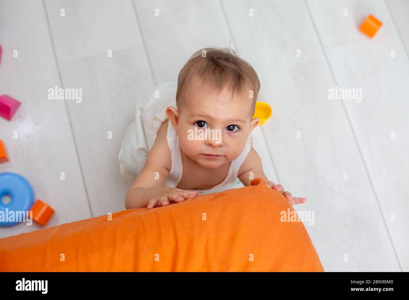 petit bébé mignon avec les yeux bruns rampent sur le coin orange du lit, essayant de se tenir sur ses pieds et regardant la caméra, vue de dessus, gros plan Banque D'Images