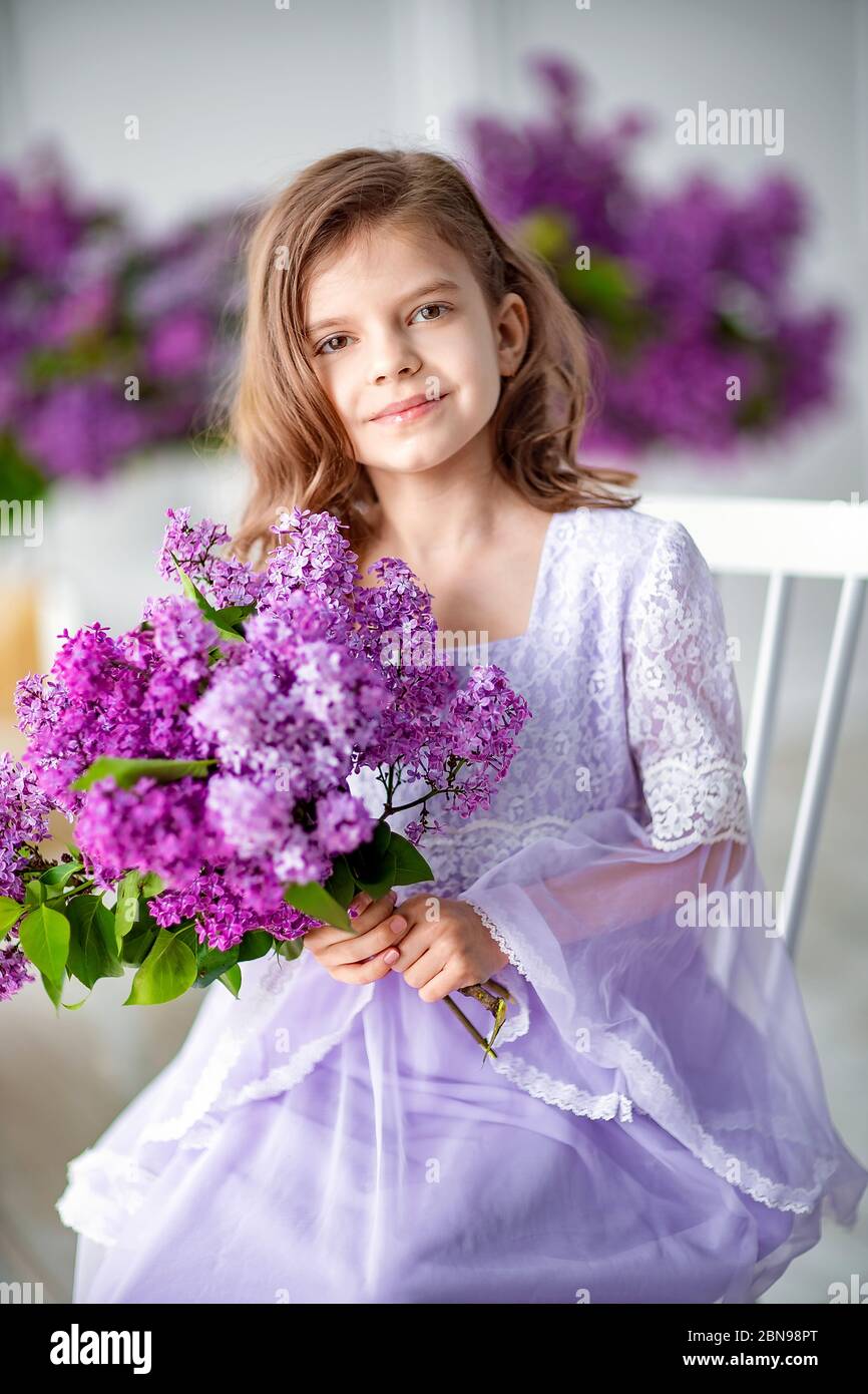 Belle petite fille d'âge préscolaire dans une délicate robe assise sur une chaise derrière balançoire décorée de fleurs de lilas. Banque D'Images