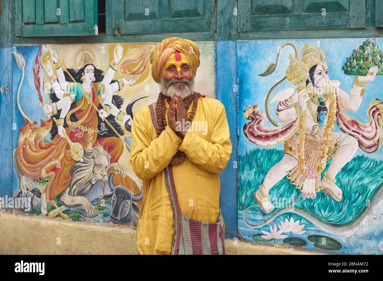 Un hindou sadhu (Saint-homme) qui exécute Namaste (salutation), Temple de Pashupatinath, Katmandou, Népal, entre les peintures murales de la déesse Durga et dieu singe Hanuman Banque D'Images