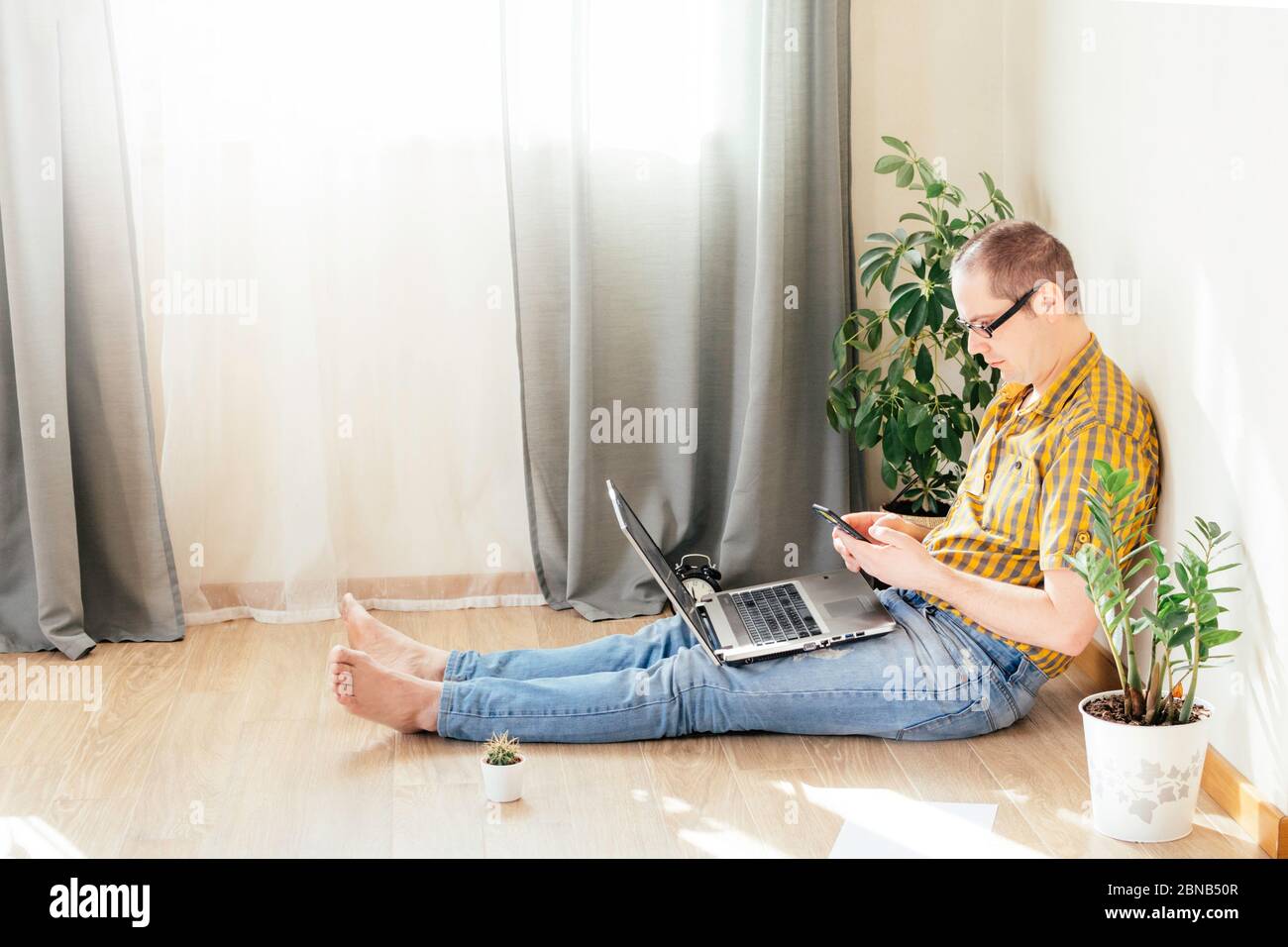 Le jeune homme est assis sur le sol et travaille à la maison sur un ordinateur avec le téléphone, des papiers et une tasse de café. Travail à distance, travail à domicile. Banque D'Images