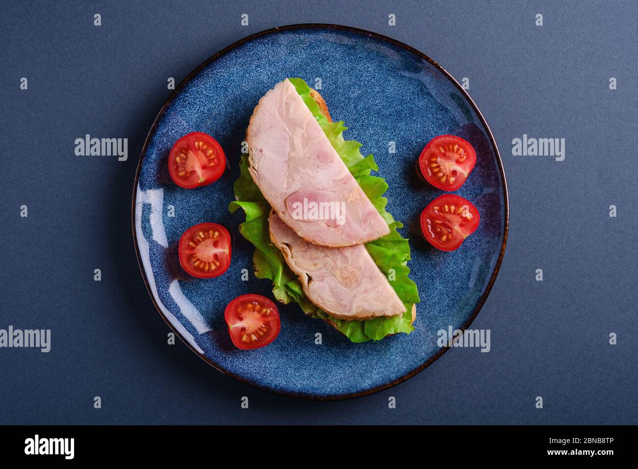 Sandwich avec viande de jambon de dinde, salade verte et tranches de tomates cerises fraîches sur une assiette bleue brillante, fond bleu minimal, vue du dessus Banque D'Images