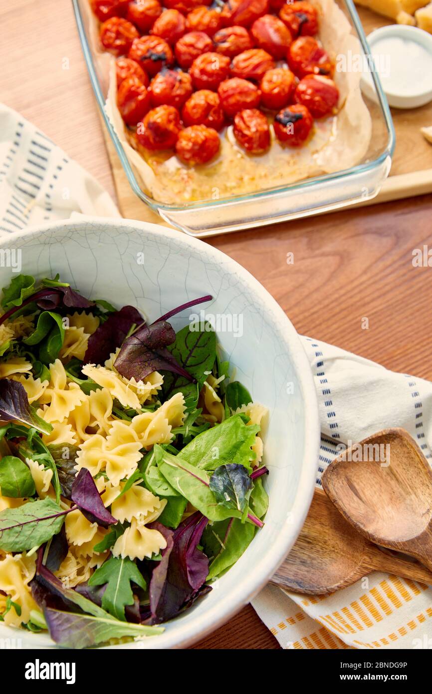 Vue en grand angle du bol avec ingrédients pour salade de pâtes près des spatules, de la serviette et du plan de coupe avec tomates dans un plat de cuisson sur fond en bois Banque D'Images