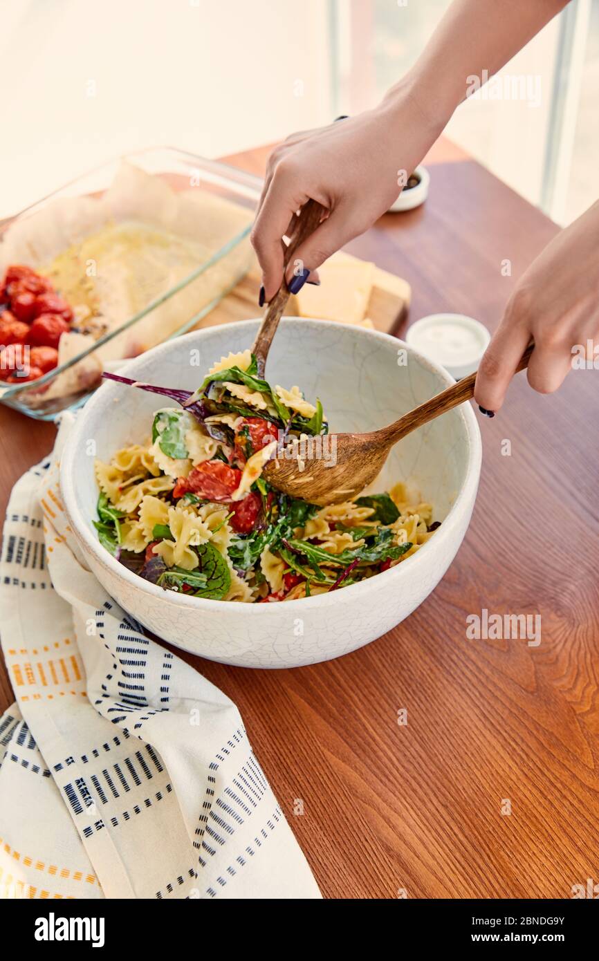 Vue courte de la femme mélangeant la salade de pâtes dans un bol avec des spatules près de la serviette à table dans la cuisine Banque D'Images