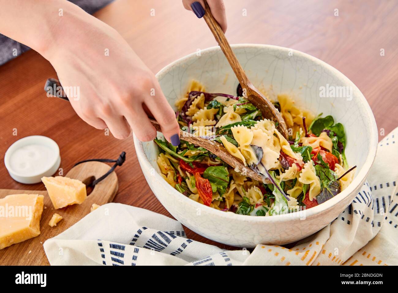 Vue partielle de la femme mélangeant la salade de pâtes dans un bol avec des spatules près de la serviette, du sel et de la planche à découper avec du parmesan près de la table dans la cuisine Banque D'Images