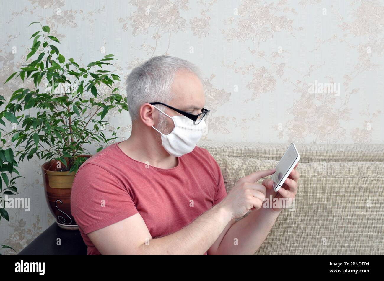 Un homme dans un masque médical, qui est à la maison sur l'auto-isolement, lit les nouvelles sur le comprimé. Concept de prévention des coronavirus Banque D'Images