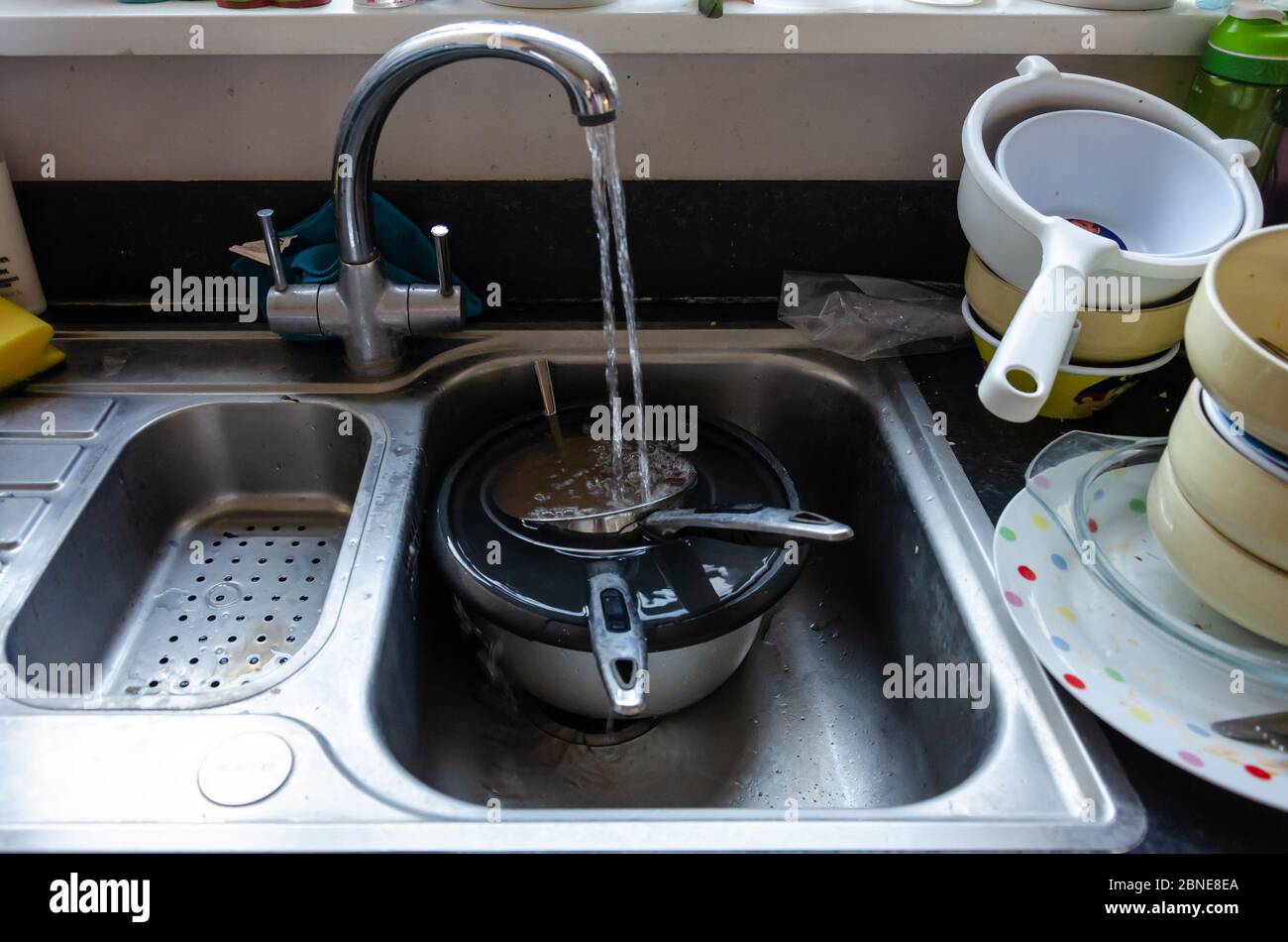 L'eau courante du robinet remplit les casseroles qui se trempent dans un évier de cuisine. Les plats sont empilés et prêts à être lavés. Banque D'Images