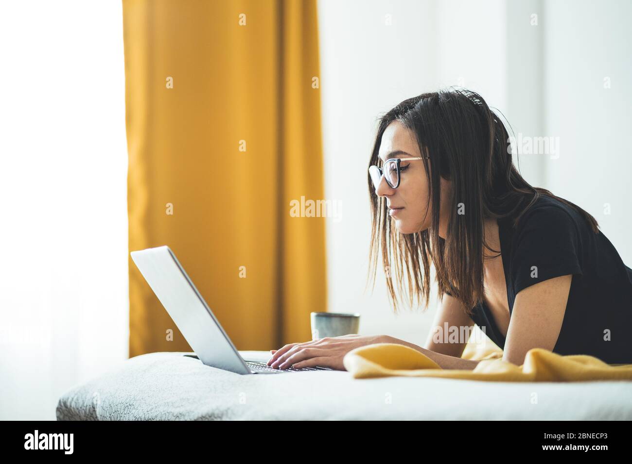 Jolie femme portant des lunettes et travaillant sur un ordinateur portable. Femme concentrée couchée au lit et travaillant à la maison. Freelance Lifestyle et corona Banque D'Images