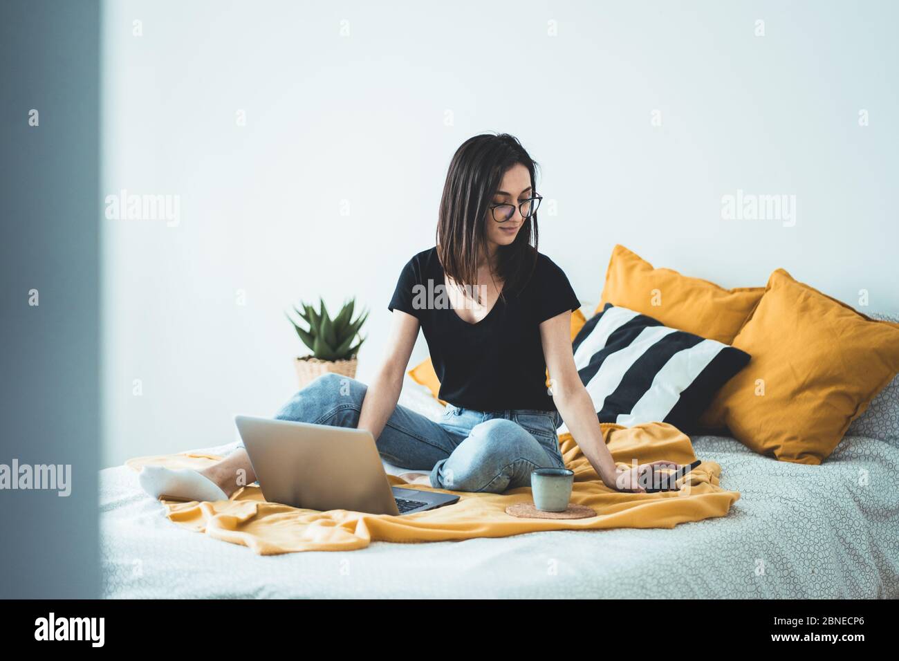 Belle jeune femme travaillant à domicile - Femme Entrepreneur assis sur le lit avec ordinateur portable, tasse à café et smartphone de chèques Banque D'Images