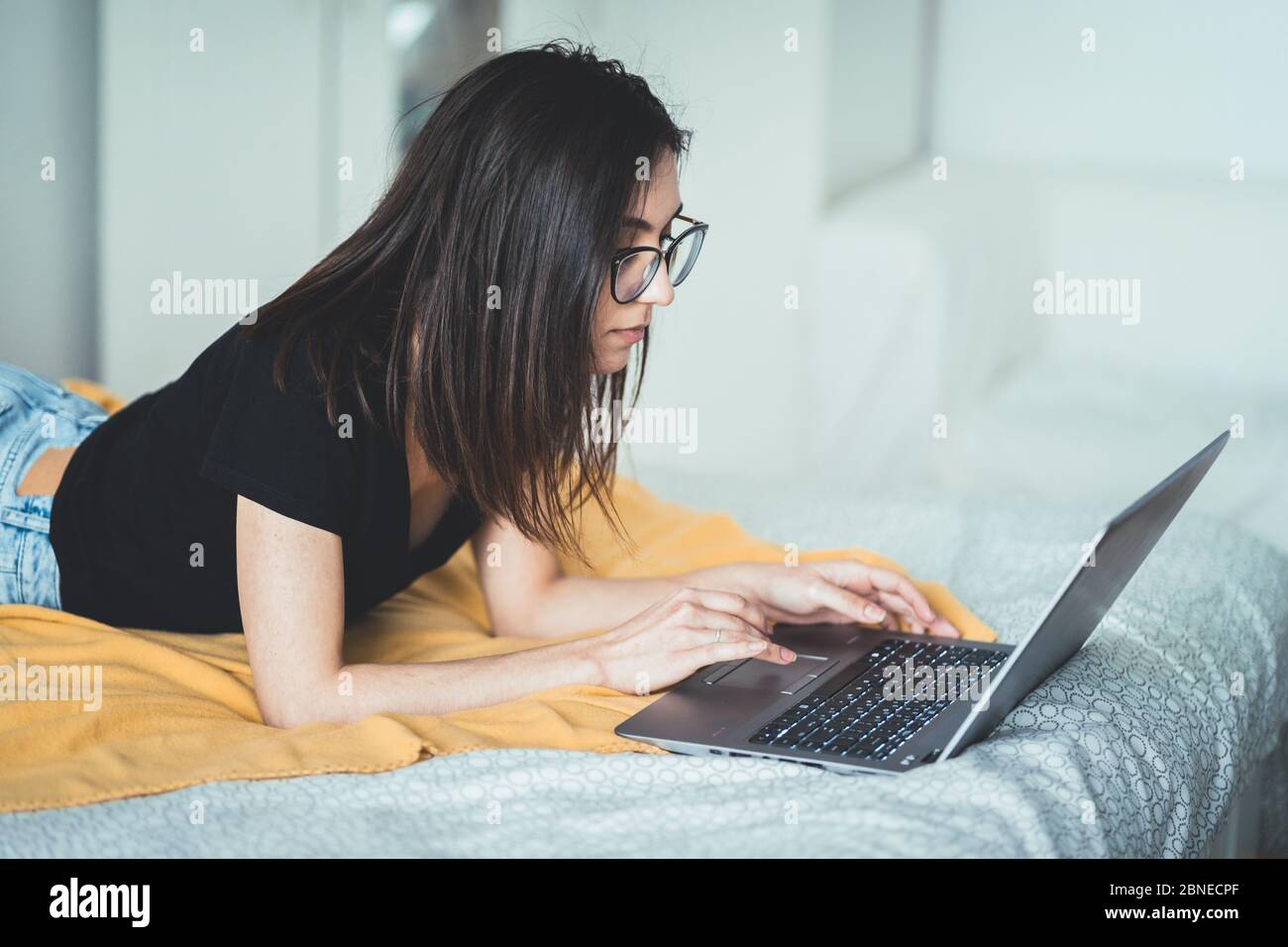 Jolie femme brune portant des lunettes et travaillant sur un ordinateur portable. Femme concentrée couchée au lit à la maison écrivant et utilisant le clavier d'ordinateur portable Banque D'Images