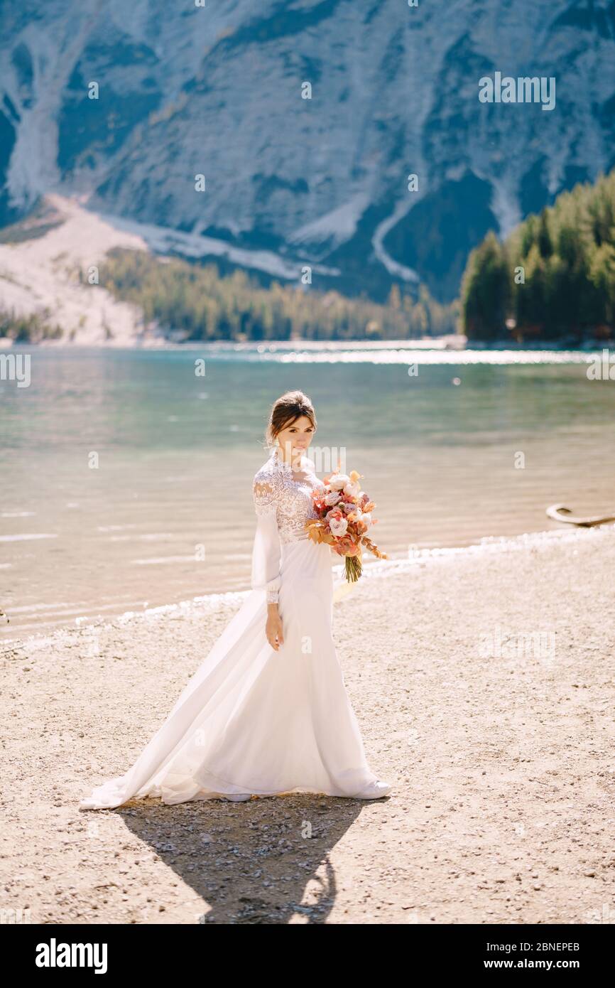 Belle mariée dans une robe blanche avec des manches et de la dentelle, avec un bouquet d'automne jaune de fleurs séchées et de roses de pivoine, sur le Lago di Braies en Italie Banque D'Images