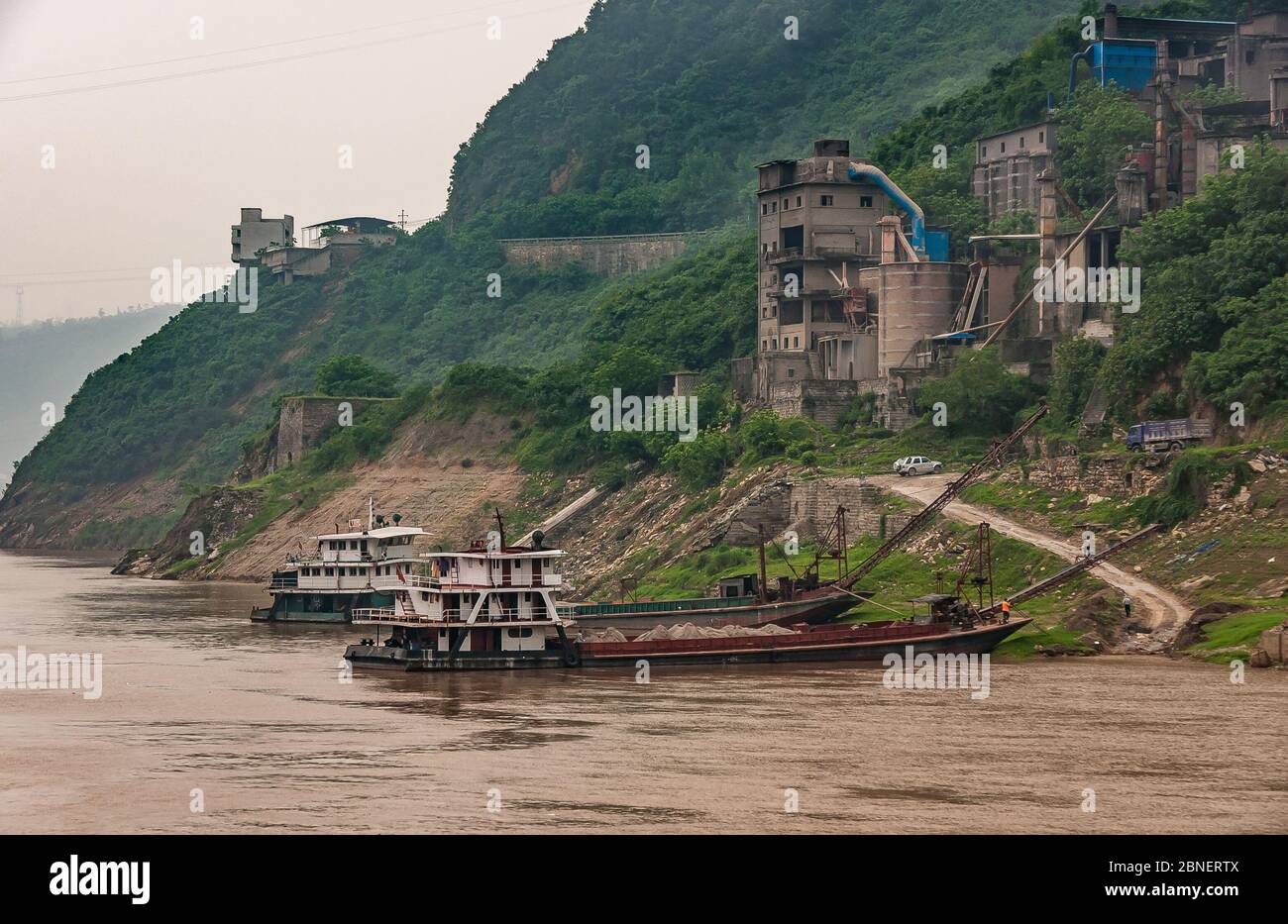 Huangqikou, Chongqing, Chine - 8 mai 2010 : fleuve Yangtze. De vieilles barges rouillées amarrées sur le rivage sous un bâtiment d'usine en pierre brune avec des ceintures mobiles t Banque D'Images