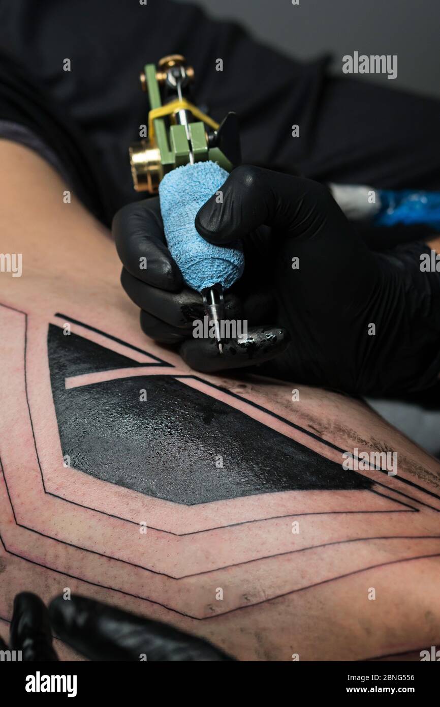 Un artiste professionnel de tatouage fait du tatouage sur la jambe de l'homme. Processus. Introduit l'encre noire dans la peau à l'aide de l'aiguille de la machine de tatouage. Gros plan sur la machine à tatouage. Tatouage. Gabarit de création d'image sur la jambe. Banque D'Images