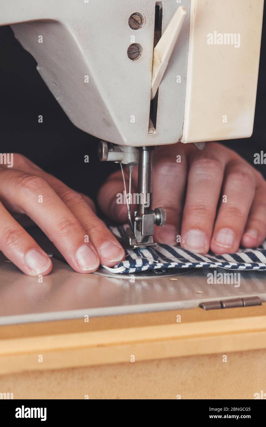 Les mains des femmes utilisant la machine à coudre pour coudre un masque médical coloré du visage pendant la pandémie de coronavirus. Masque de protection fait maison contre le virus. Masques à coudre, COVID-19. Mise au point sélective. Banque D'Images