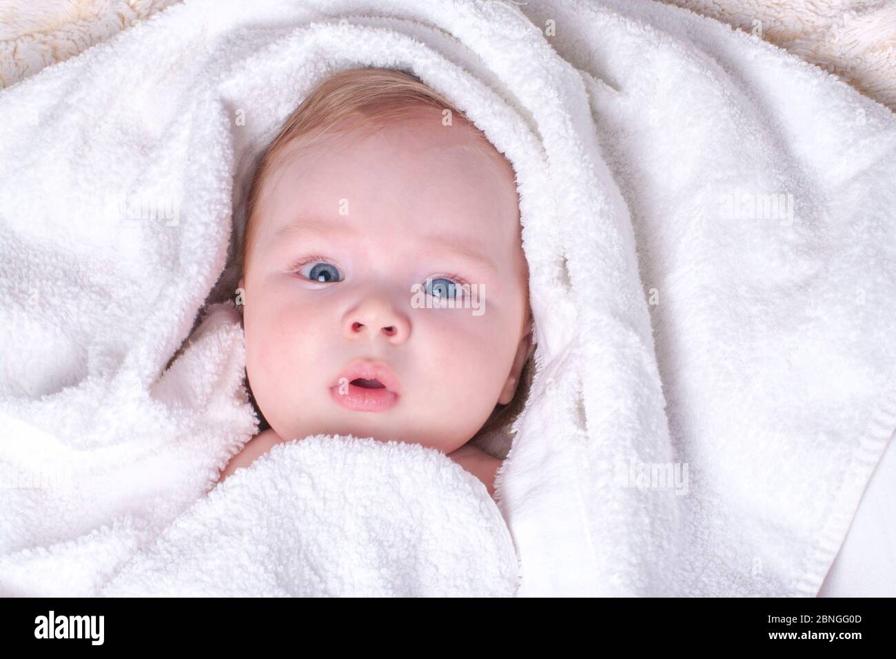 Charmant, surpris, bleu-eyed bébé avec des lèvres et des cheveux blonds se  trouve dans une serviette sèche, blanche après avoir baigné dans la salle  de bains. Concept de soins de la peau,