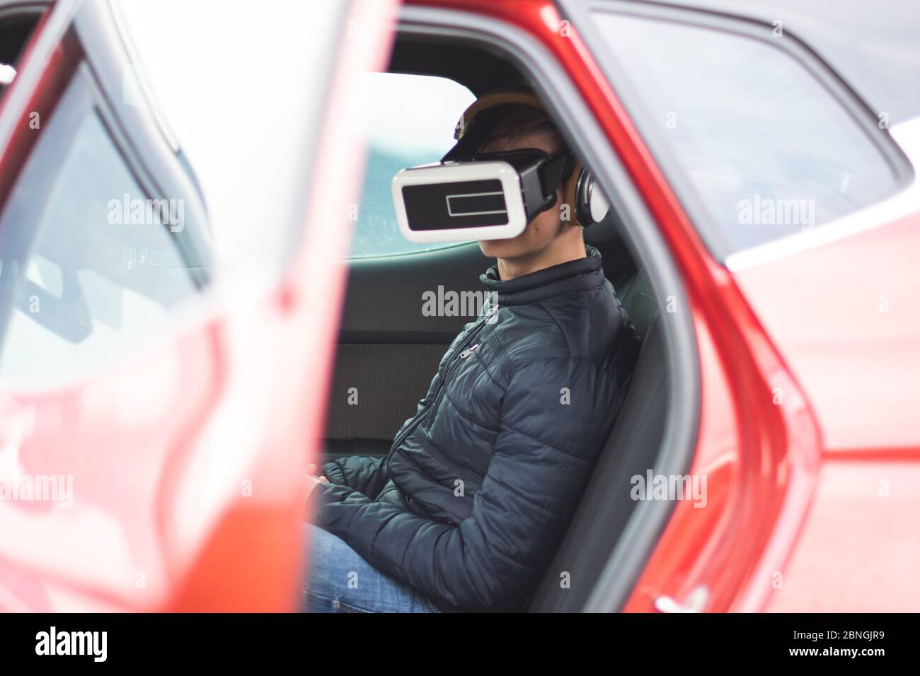 Vue d'un homme utilisant la réalité virtuelle pendant qu'il est assis le siège arrière d'une voiture rouge avec sa porte ouvrir Banque D'Images