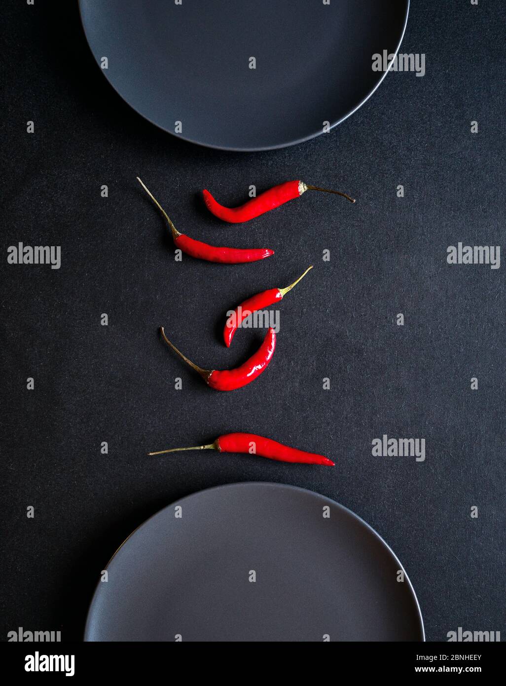 Composition minimaliste de piments rouges entre deux assiettes en céramique sur table noire Banque D'Images