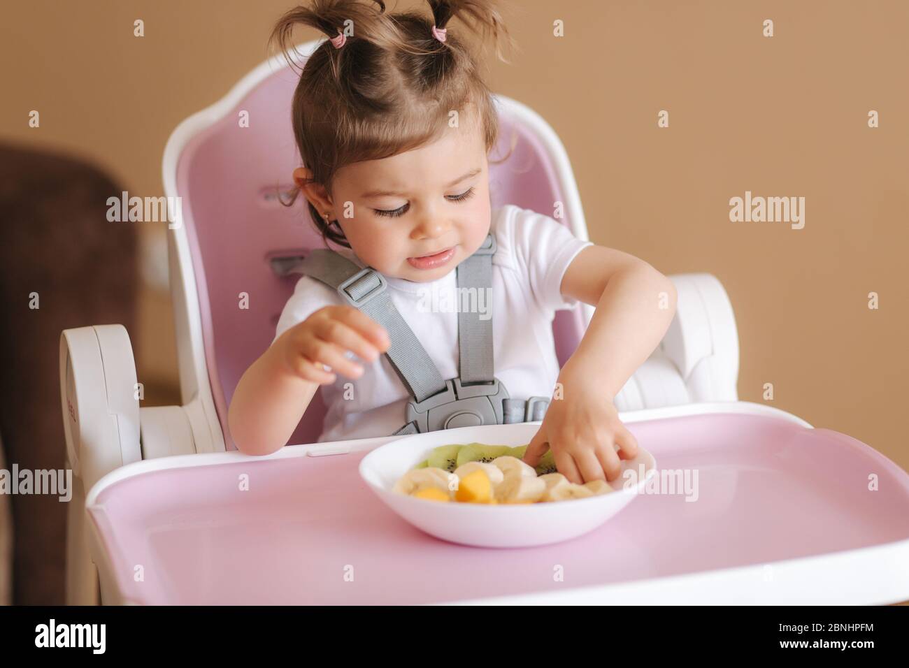 Belle petite fille de bébé la première fois manger des fruits exotiques  dans la chaise haute. La petite fille mignonne a un goût délice de fruist,  de mangue, de kiwi et de