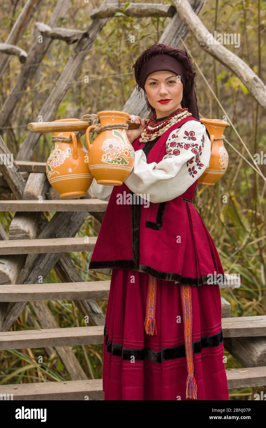 Une femme en vêtements traditionnels transporte de l'eau dans de vieux  barils. Les vieux carafes sont portés par une fille dans un costume  stylisé. Des vêtements originaux et magnifiques Photo Stock -