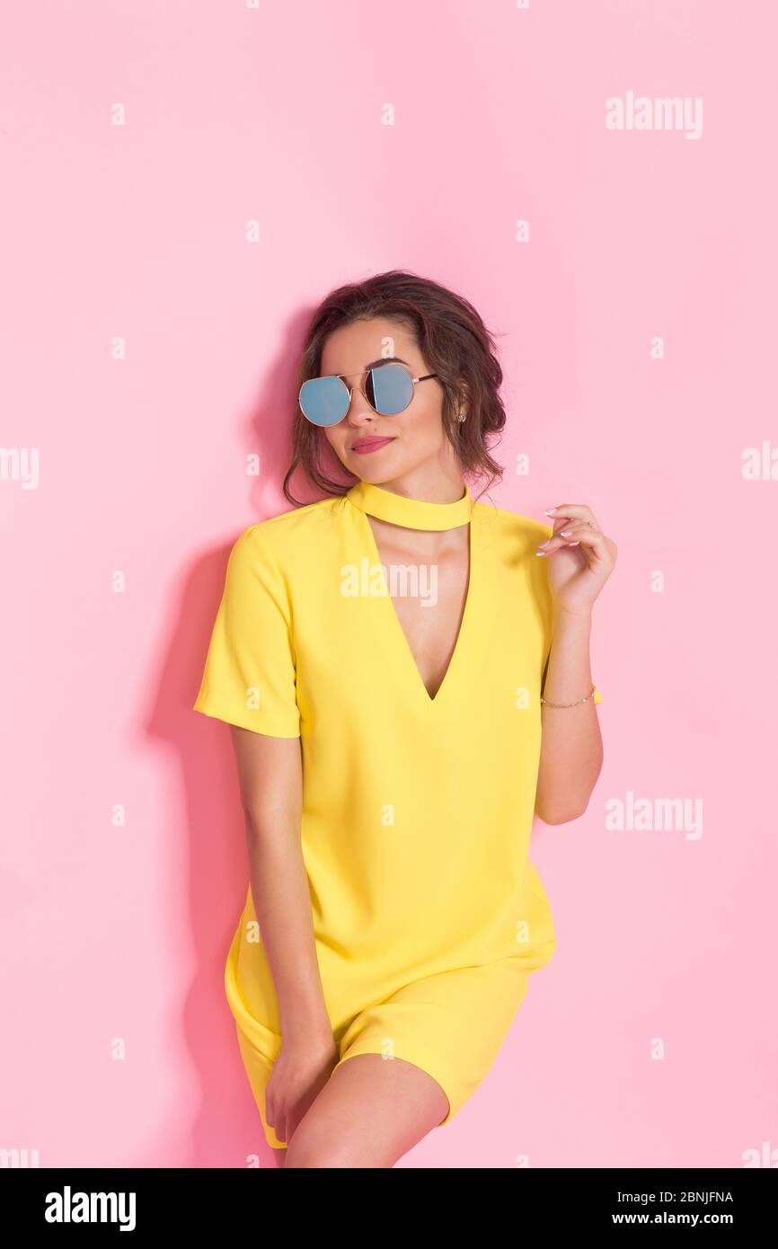 Belle fille portant une robe jaune et des lunettes de soleil posant sur fond rose en studio. Banque D'Images