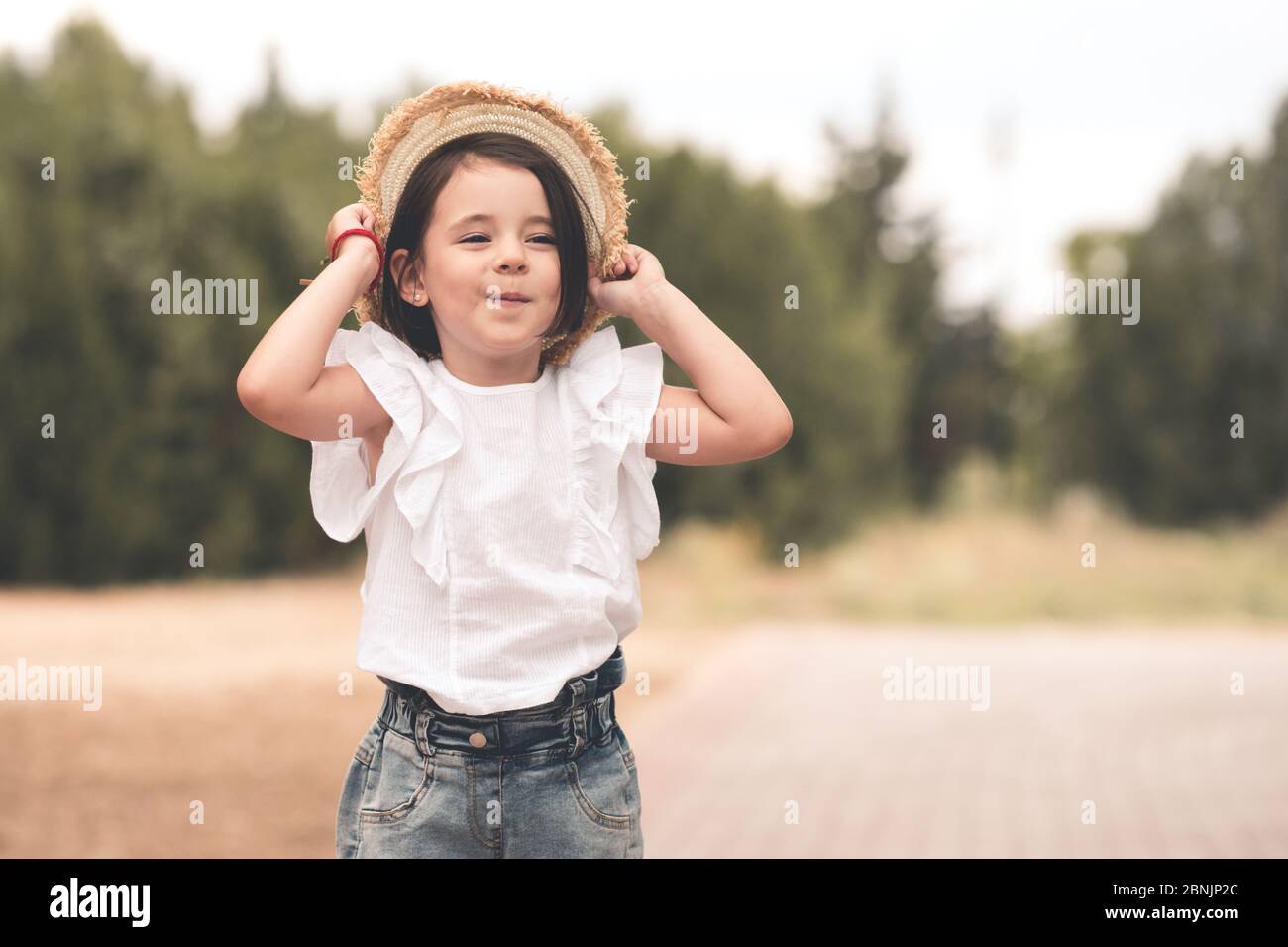 Drôle mignon enfant fille 3-4 ans tenant chapeau de paille portant le  dessus blanc et short en denim posant dans le parc sur fond de nature.  Bonheur. Assaisonnement d'été Photo Stock -