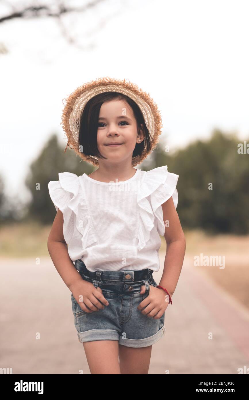 Jeune fille souriante et tendance 4-5 ans, vêtue d'un chapeau de paille et d'un haut blanc d'été avec short en denim à l'extérieur sur fond de nature. Banque D'Images