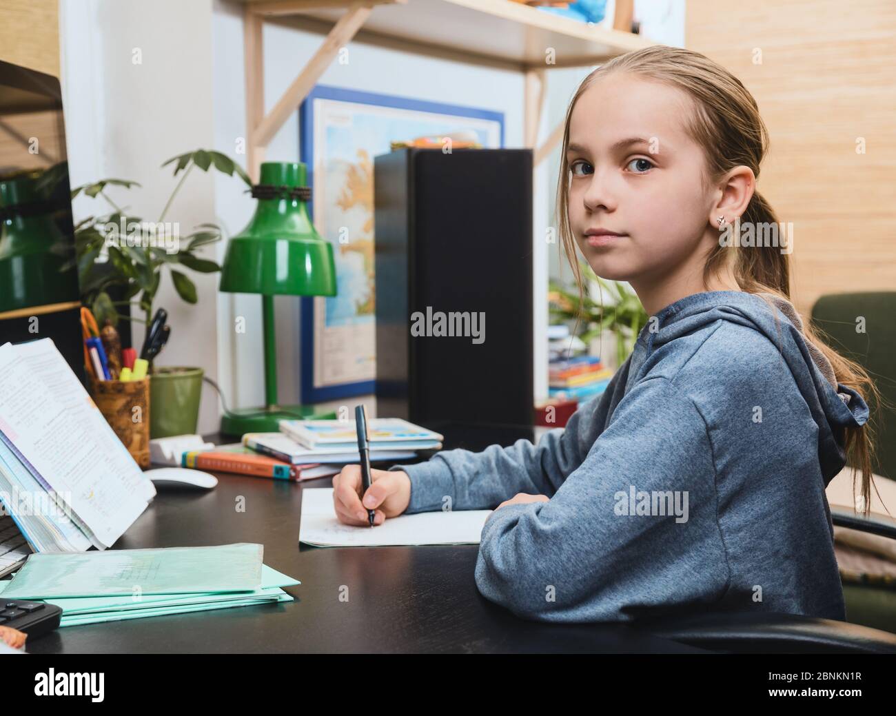 Écolière écrivant dans un carnet assis près d'une table. Elle étudie à la maison pour faire ses devoirs pendant son auto-isolement. Concept de homeschooling Banque D'Images