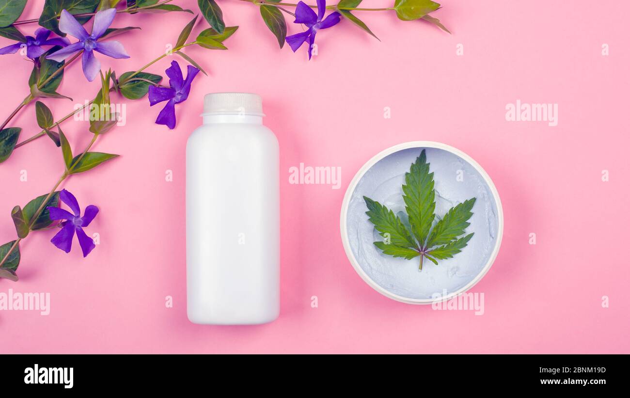 bouteilles blanches avec cosmétiques sur fond rose avec une feuille de marijuana et des fleurs sauvages violettes vue du dessus. beauté, soins de la peau. Banque D'Images