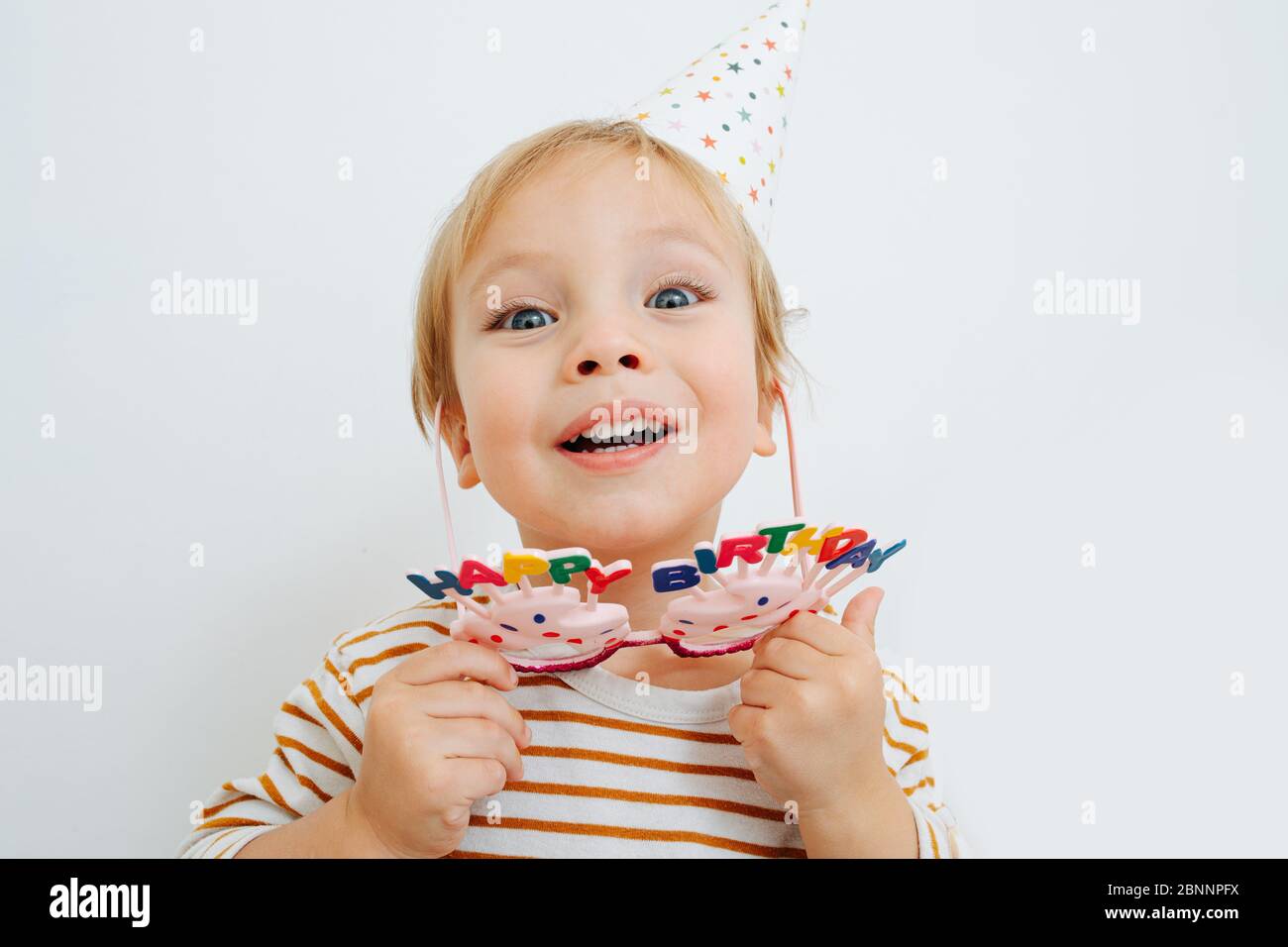 Mignon enfant prenant des verres d'anniversaire heureux et chapeau de fête conique sur blanc Banque D'Images