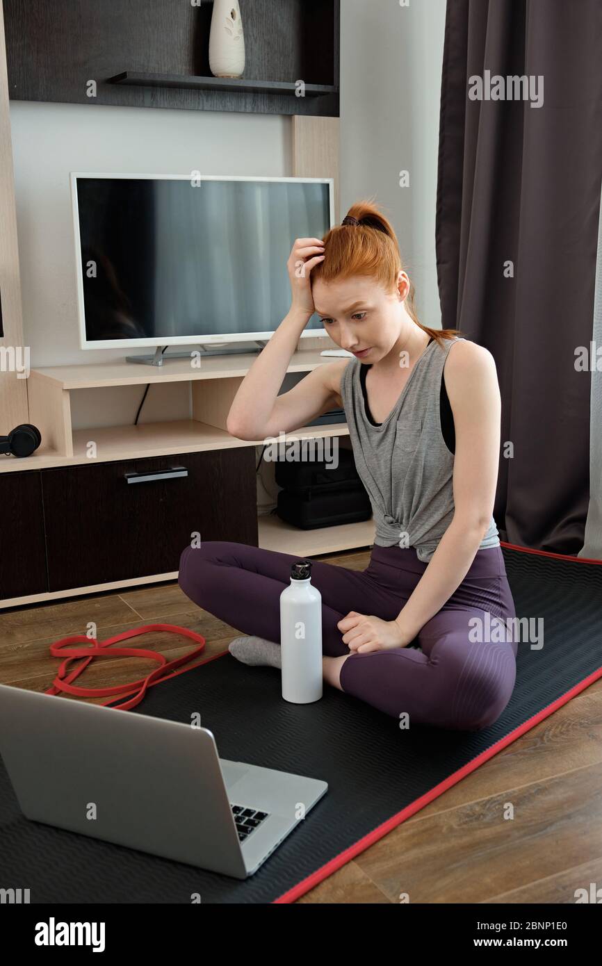 La jeune fille aux cheveux rouges est assise sur un tapis de sport dans un uniforme d'entraînement, levant les sourcils et égratignures de tête, regardant l'ordinateur portable Banque D'Images