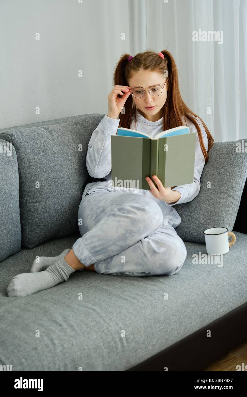 Une fille de pyjama aux cheveux rouges est assise sur le canapé avec ses jambes repliées et lit un livre, en ajustant ses lunettes Banque D'Images