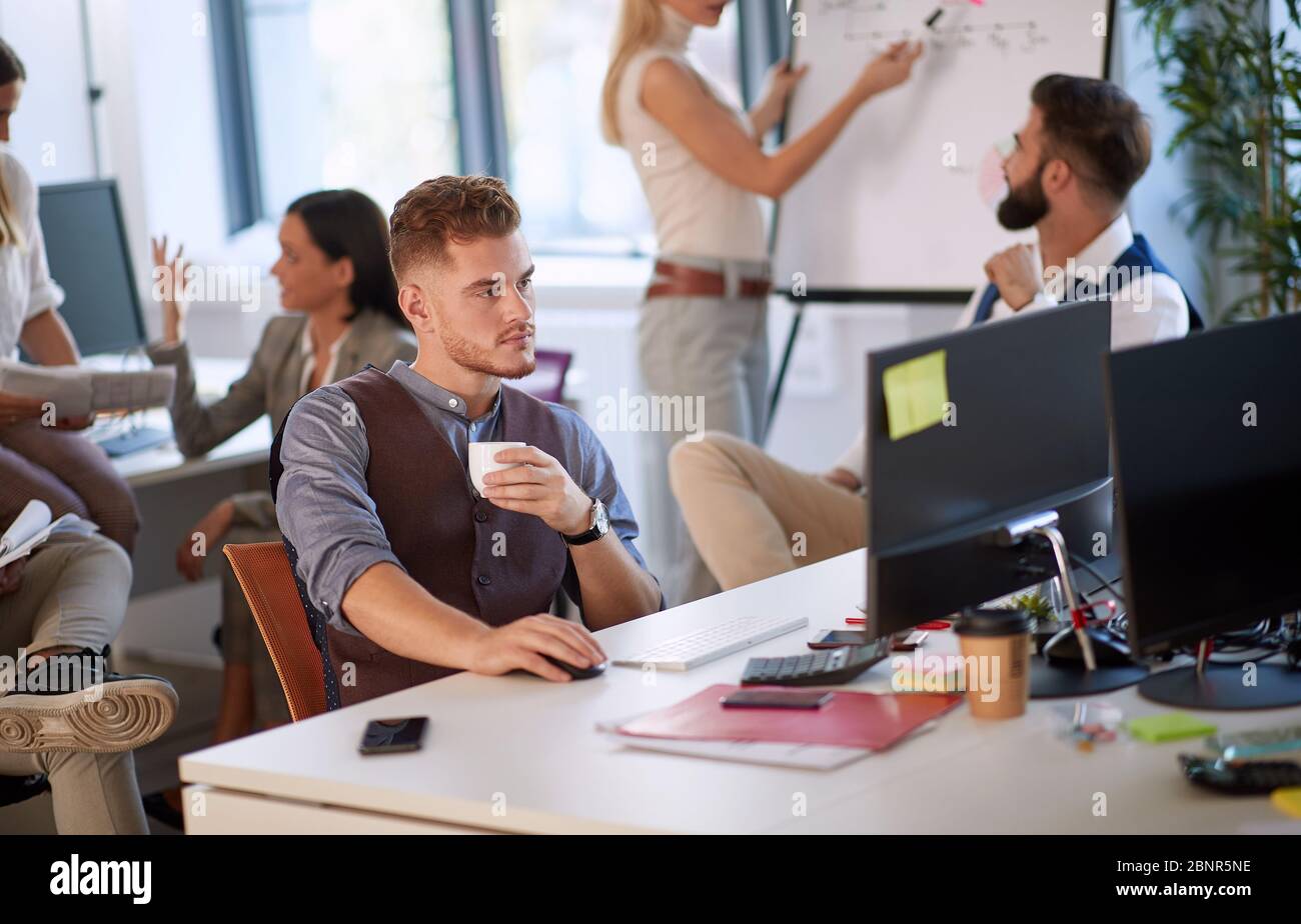 Jeune entrepreneur tenant une tasse de café tout en étant assis et travaillant dans le bureau plein de personnes Banque D'Images