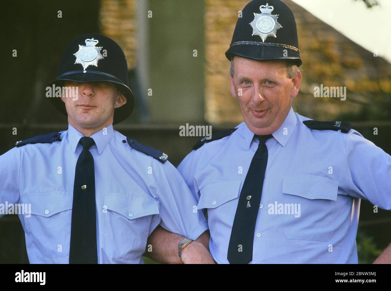 Deux gendarmes de police masculins se posent pour rire avec des casques de gardien de taille incorrecte. Angleterre, Royaume-Uni. Vers les années 1980 Banque D'Images