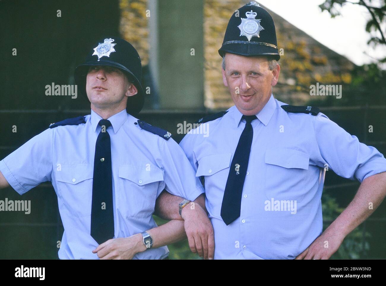 Deux gendarmes de police masculins se posent pour rire avec des casques de gardien de taille incorrecte. Angleterre, Royaume-Uni. Vers les années 1980 Banque D'Images