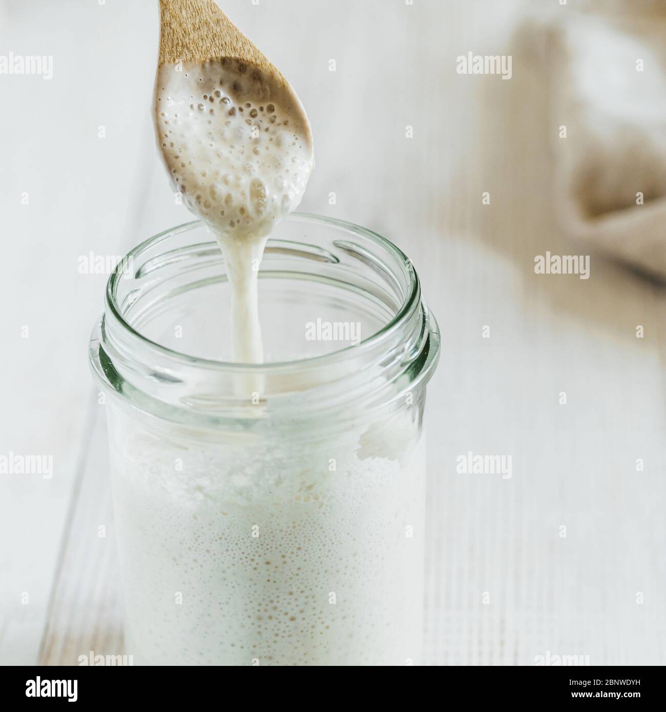Prêt à préparer du pain de pâte de blé de démarrage goutte à goutte à partir d'une cuillère en bois. Vue rapprochée du bol en verre avec démarreur à la levain hydratation plus élevée sur fond en bois blanc. Concept de pain maison Banque D'Images