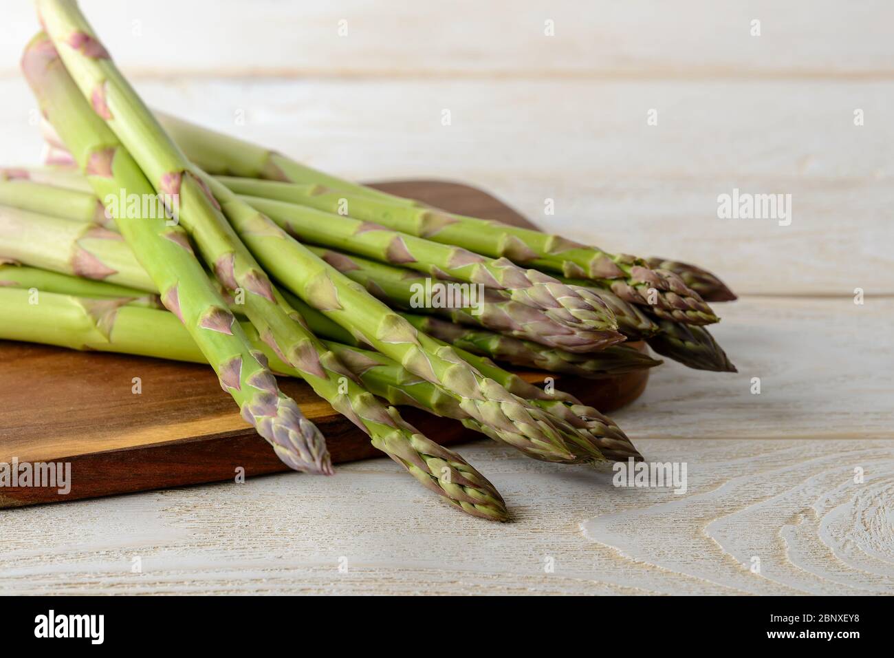 Tiges d'asperges vertes fraîches crues sur une planche à découper brune sur une table de cuisine en bois blanc. Ingrédient pour manger de la viande, régime végétarien et alimentaire cru. Banque D'Images